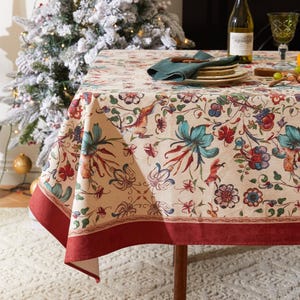 May include: A cream-coloured tablecloth featuring a floral and animal design in red, blue, and green. The tablecloth has a dark red border and is set on a wooden table. A Christmas tree is in the background.