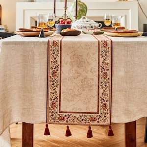 May include: A beige table runner with a floral pattern and red tassels. The runner has a border with a repeating pattern of pumpkins, leaves, and flowers.