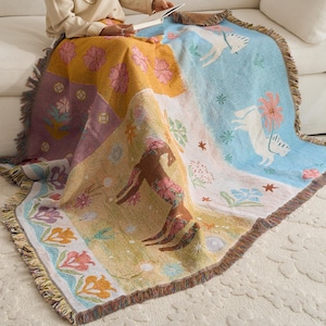 Könnte beinhalten: Eine gewebte Wohndecke mit einem mehrteiligen Design, bestehend aus einem hellblauen Bereich mit weißen Pferden und rosa Blumen, einem gelben Bereich mit einem braunen Pferd und einem violetten Bereich mit Blumenmustern, abgeschlossen mit Fransen.