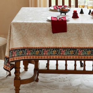 Pode incluir: Uma toalha de mesa bege com um padrão floral e uma borda decorativa. A borda apresenta um design repetido de flores e formas geométricas em tons de rosa, verde e laranja. A toalha de mesa está sobre uma mesa de madeira.