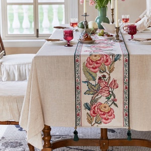 May include: A beige linen tablecloth with a floral patterned table runner. The table runner features pink and green flowers with a green border. The table is set with glasses and a plate.