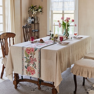 Pode incluir: Uma toalha de mesa de linho bege com um corredor bordado com flores. O corredor apresenta rosas rosa e vermelhas, folhas verdes e uma borda verde escura. A mesa está posta com duas cadeiras, castiçais e copos.