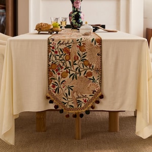 May include: A cream tablecloth with a decorative table runner. The runner features a detailed pattern of birds, pomegranates, and foliage in shades of beige, red, and green. The runner is trimmed with a gold border and tassels.
