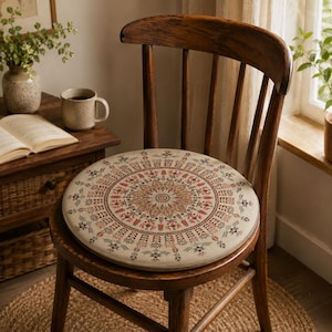 Könnte beinhalten: Ein rundes, beiges Sitzkissen mit einem detaillierten, symmetrischen Mandala-Muster in Rot-, Schwarz- und gedämpften Grüntönen. Das Kissen liegt auf einem dunkelbraunen Holzstuhl mit geschwungener Rückenlehne und vertikalen Stäben.