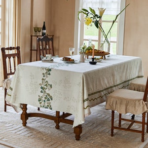 May include: A white tablecloth with a green floral pattern, set on a wooden table with four chairs. The tablecloth has a green border with a scalloped edge. The table is set with a vase of yellow flowers, a bowl of fruit, and a plate with a cup and saucer.