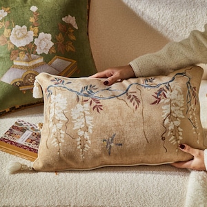 May include: A beige throw pillow with a white floral design and a blue vine pattern. The pillow has a white border and a tassel.