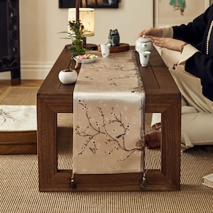 Può includere: Un tavolo di legno con un runner a motivi floreali e due tazze da tè. Il tavolo è apparecchiato per una cerimonia del tè con una teiera e una piccola pianta in un vaso bianco.