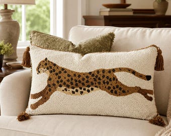 May include: A rectangular throw pillow with a cream-colored base and a textured, hooked-wool design of a running cheetah in shades of brown and black. The pillow features brown tassels at each of the four corners and a soft, tufted surface texture.