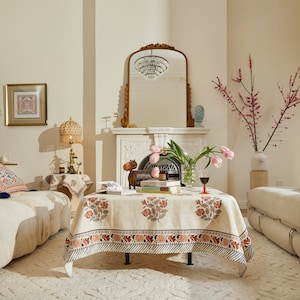 May include: A white tablecloth with a floral print in shades of pink and brown, covering a square table with a black metal base. The table is set with a glass of red wine and a stack of books. A wooden figurine of a hippopotamus sits on the table.