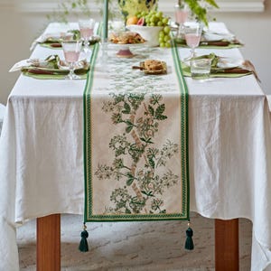 Peut inclure: Une nappe blanche avec un chemin de table floral vert et beige. Le chemin de table présente un motif botanique et des glands verts. La table est dressée avec des assiettes, des verres et des couverts, suggérant un cadre de repas.