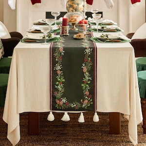 May include: A festive dining table set for a holiday meal. The table is covered with a cream-colored tablecloth and a dark green runner with floral and berry embroidery. The table is set with plates, glasses, and candles.