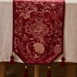 Puede incluir: Camino de mesa burdeos con un detallado diseño floral y de jarrón en crema. El corredor tiene una textura de terciopelo y está adornado con borlas en cada extremo, agregando un toque de elegancia a cualquier mesa.