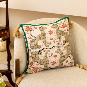 May include: A decorative cushion with a green border and a white background featuring a repeating pattern of orange flowers and brown leopards. The cushion has a tassel on each corner.