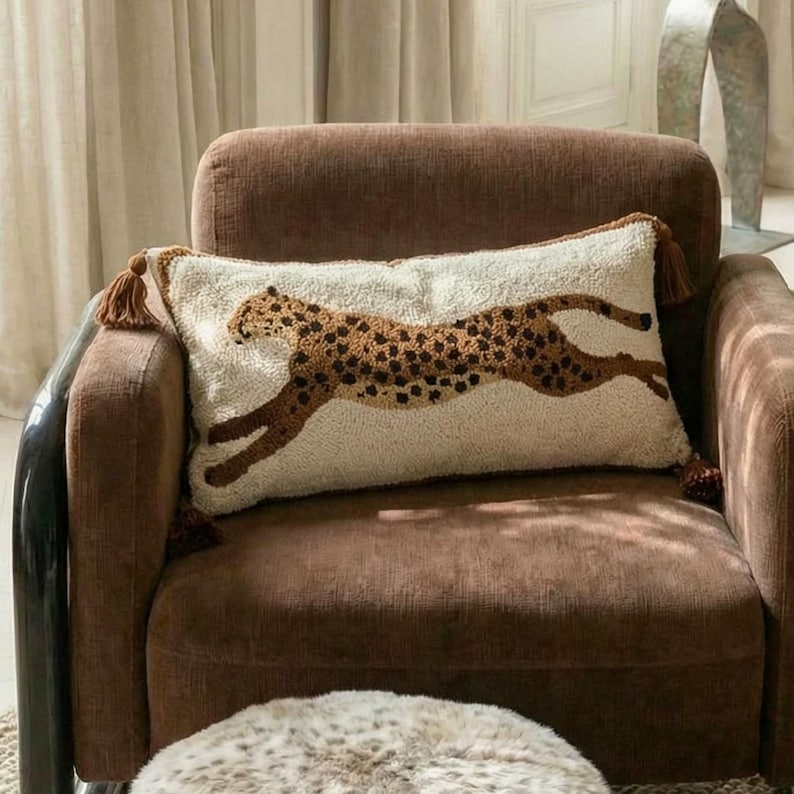 Puede incluir: Un coj&iacute;n rectangular con una superficie texturizada de color crema que presenta un gr&aacute;fico de leopardo marr&oacute;n y negro. El coj&iacute;n tiene borlas marrones en cada una de las cuatro esquinas y est&aacute; colocado sobre un sill&oacute;n tapizado de color marr&oacute;n.