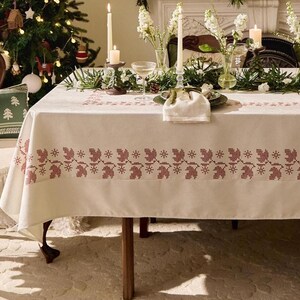 May include: A cream tablecloth with a red embroidered border, featuring a repeating pattern of figures. The table is set with candles, flowers, and glassware, creating a festive atmosphere.
