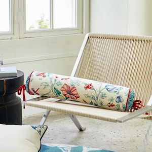May include: A white cylindrical cushion with a floral pattern in shades of red, blue, and green. The cushion has red and blue trim and is sitting on a white wicker chair.
