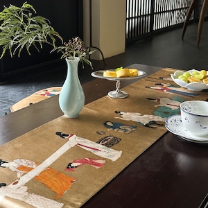 May include: A gold table runner featuring a colourful print of women in traditional dress. The runner is displayed on a dark wood table with a light blue vase, a silver serving tray with small cakes, and a white teacup and saucer.