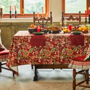 Puede incluir: Un mantel de mesa rojo floral con detalles dorados, colocado sobre una mesa de madera con un centro de mesa de fruta y velas. La mesa está decorada con servilletas rojas y platos verdes.