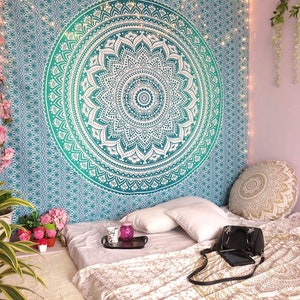 May include: A teal and white mandala tapestry hangs on a wall above a bed with white pillows and a gold and white patterned throw blanket. A black purse and a tray with two mugs and a glass are on the bed.