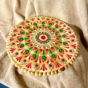 May include: A wooden round cake stand with a hand-painted mandala design in red, green, and white. The stand has a beaded edge.