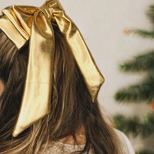 May include: A large gold bow made of satin fabric is tied in a woman's hair.