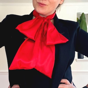 May include: A bright red satin neck scarf tied in a large bow, worn over a black blazer. The scarf is a vibrant contrast against the dark jacket, creating a bold and stylish look. The scarf's smooth texture and rich color are prominent.