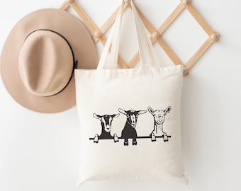 May include: A white canvas tote bag with three black and white goats peeking over the top. The goats are looking at the camera. The bag is hanging on a wooden rack.