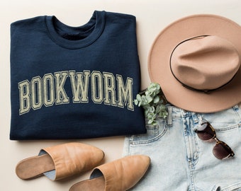 Pode incluir: Uma camisa de moletom azul marinho com o texto "BOOKWORM" em uma fonte de estilo universitário vintage. A camisa de moletom está dobrada e colocada sobre uma superfície bege claro. Também são mostrados um par de tamancos de couro marrom, um par de shorts jeans azul claro, um chapéu fedora marrom e um par de óculos de sol marrons.