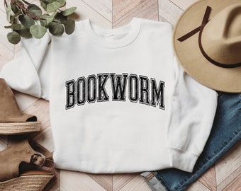 Pode incluir: Camisola branca com o texto "BOOKWORM" em preto, em uma fonte de estilo vintage desgastada. A camisola está colocada sobre um fundo de madeira com um par de cunhas marrons e um chapéu fedora marrom.