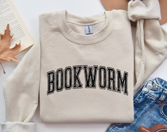 Pode incluir: Uma camisola bege com a palavra "BOOKWORM" impressa a preto na frente. A camisola está dobrada e colocada sobre uma superfície branca com um livro, um chapéu e calças de ganga.