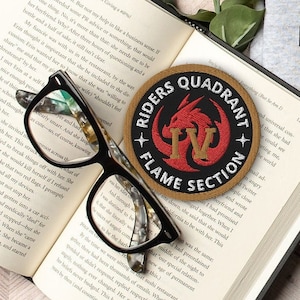 May include: A brown and tan patch with a red dragon and the text "Riders Quadrant Flame Section" on a book with a pair of black glasses. The book is open to a page with text about the "Subtle Art of Not Giving a F***".
