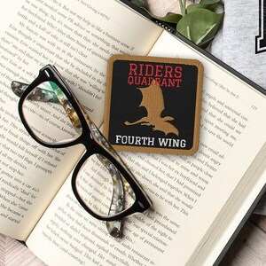 May include: A brown leather patch with the text "Riders Quadrant Fourth Wing" and a black silhouette of a bird. The patch is on a book with a pair of glasses on top.