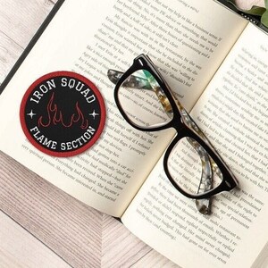 May include: A black and red embroidered patch with the text "Iron Squad Flame Section" and a stylized flame design. The patch is lying on an open book with a pair of black framed glasses.