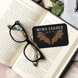 May include: A black and gold embroidered patch with the text "WING LEADER" and two stylized wings. The patch is on a book with a pair of reading glasses.
