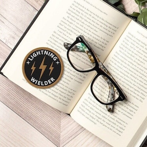 May include: An open book with black-framed eyeglasses resting on the pages. A round patch with the text "LIGHTNING WIELDER" and lightning bolt graphics is on the left. A gray sweatshirt with the word "BA" is visible in the background.