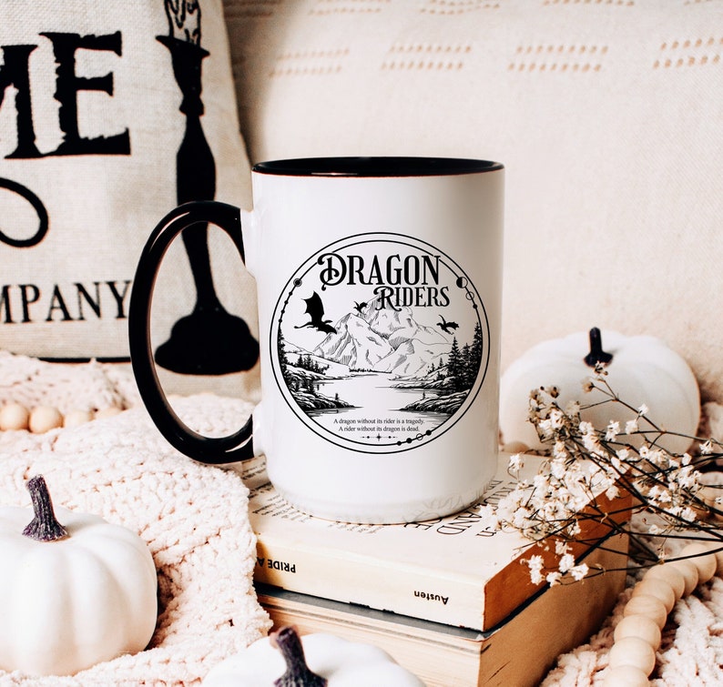 May include: White ceramic mug with a black rim and handle. The mug features a black and white illustration of a dragon and mountains with the text "Dragon Riders" in a circle. The quote "A dragon without its rider is a tragedy. A rider without a dragon is in exile." is printed below the illustration.