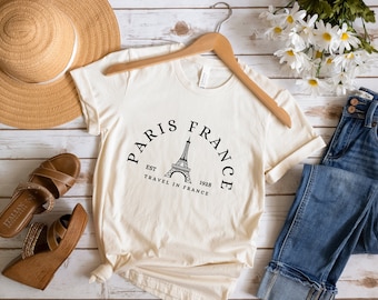 Op de afbeelding: Een wit T-shirt met een zwarte afbeelding van de Eiffeltoren en de tekst "Paris France Est 1928 Travel in France".