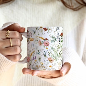 May include: White ceramic mug with a floral pattern featuring pink, yellow, and blue flowers, green leaves, and brown stems on a white background.