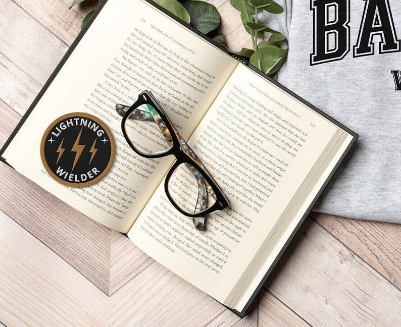 May include: An open book with black-framed spectacles on the pages. A round patch with the text "LIGHTNING WIELDER" and lightning bolt graphics is on the left. A grey sweatshirt with the word "BA" is visible in the background.