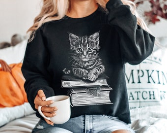 Op de afbeelding: Zwart sweatshirt met een grafische afbeelding van een kat die op een stapel boeken zit. De kat is in zwart-wit getekend.