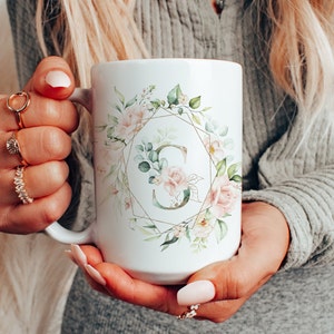 May include: White ceramic mug with a floral design and a gold geometric frame. The letter "S" is in the center of the frame.
