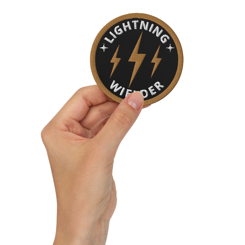 May include: Circular embroidered patch with a black central design of three gold lightning bolts and the words "LIGHTNING WIFDER" in white. The patch has a gold border and white stars. The patch is held in a hand.