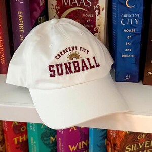 May include: A white baseball cap featuring the embroidered text "CRESCENT CITY SUNBALL" in burgundy. The cap has a curved peak and a traditional six-panel construction. The text is arched above a sunburst graphic.