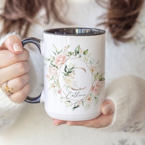 May include: White ceramic mug with a black rim and handle. The mug features a watercolor design of pink and white flowers, green leaves, and a gold geometric frame with the letter "C" in the center. The name "Catherine" is written below the design.