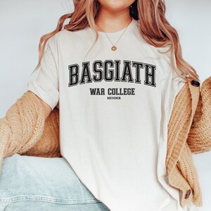 Op de afbeelding: Een wit T-shirt met zwarte tekst die "BASGIATH WAR COLLEGE MENDER" luidt. De tekst is in een college-lettertype.
