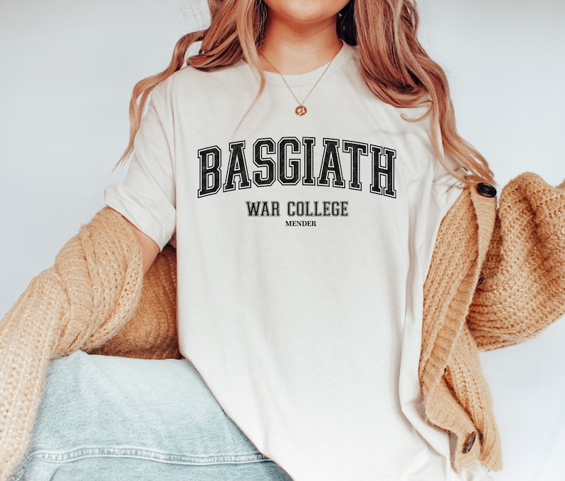 Op de afbeelding: Een wit T-shirt met zwarte tekst die "BASGIATH WAR COLLEGE MENDER" luidt. De tekst is in een college-lettertype.