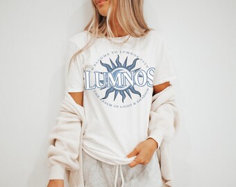 May include: White t-shirt with a blue graphic design featuring a sun with rays and the text "Welcome to Lumnos City" and "Lumnos The Realm of Light & Shadow".