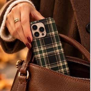 May include: A smartphone case featuring a dark green, black, and beige plaid pattern. The case has a rectangular cutout for the camera lenses and is shown being placed into a brown textured leather handbag with gold-toned hardware accents.