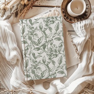 May include: A white journal with a green floral pattern. The journal is lying on a white surface with a brown and white blanket.