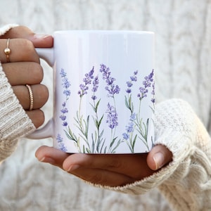 May include: White ceramic mug with a lavender floral design. The mug is being held by a person wearing a white sweater.